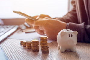 Man holding phone sitting next to white piggy bank and stacked coins